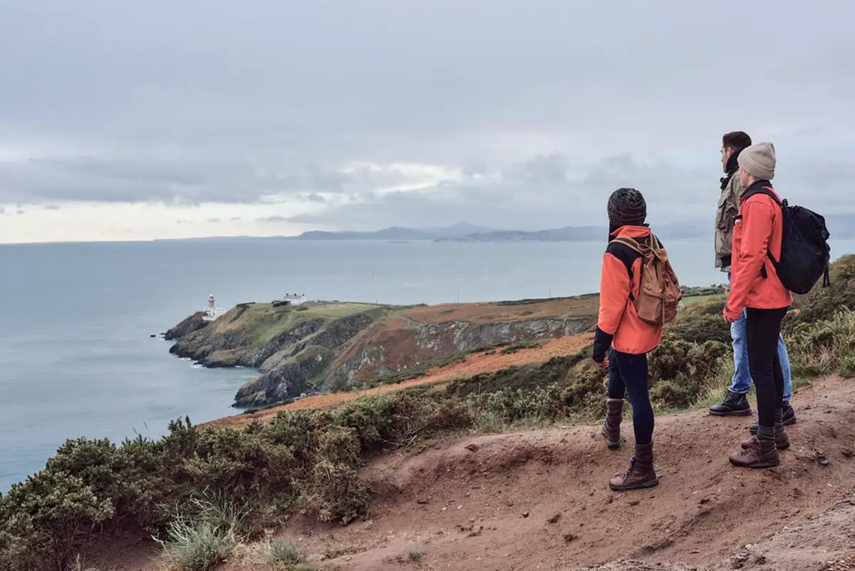 Three walkers admire the view of the lighthouse from Howth Cliff walk in Dublin.