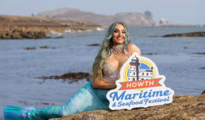 Mermaid on the rocks, smiling, holding a sign for Maritime & Seafood Festival.