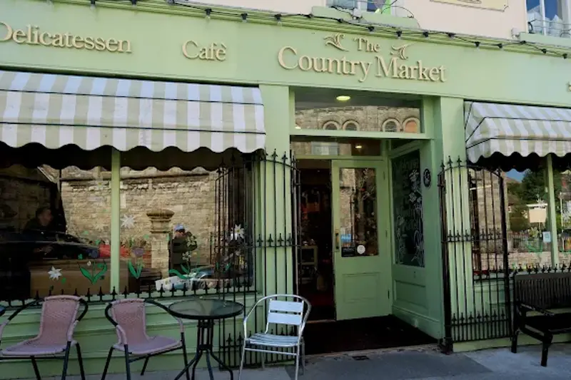 Shopfront of 'The Country Market' in Howth, with pale green sign, beige lettering and tables and chairs outside.