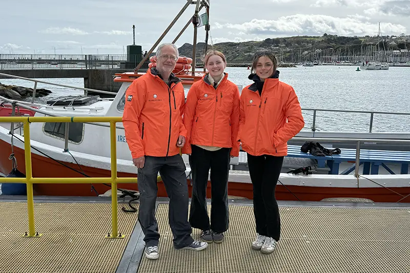 Three people wearing matching orange jackets in front of ferry on Howth Pier.